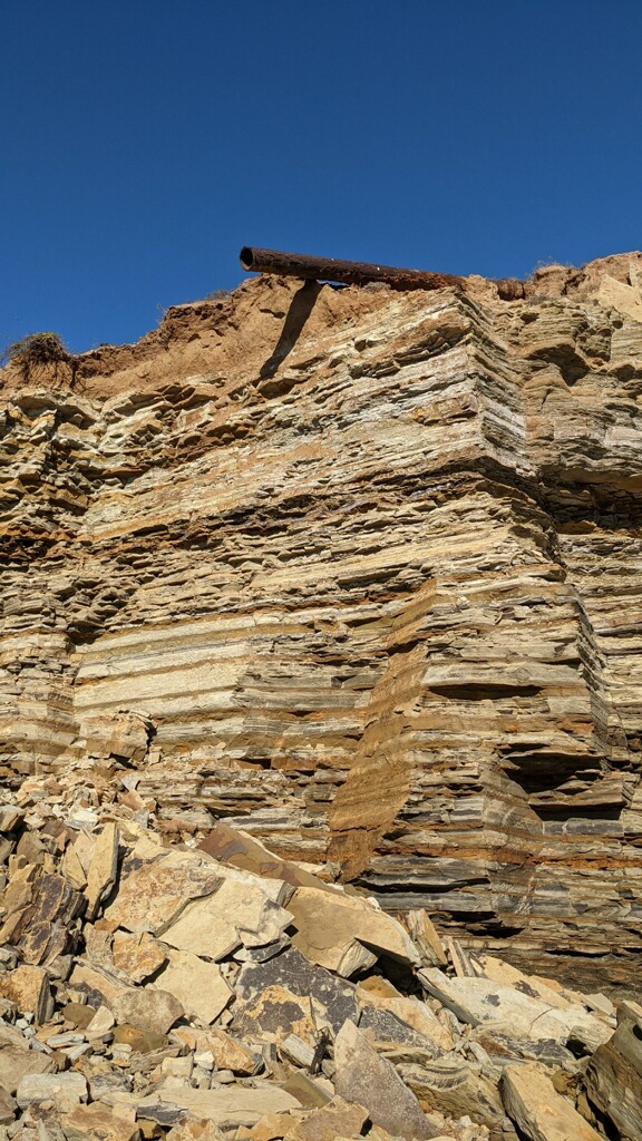 Broken sandstone rocks against a layered cliff. A rusted drainage pipe sits of top of the cliff.