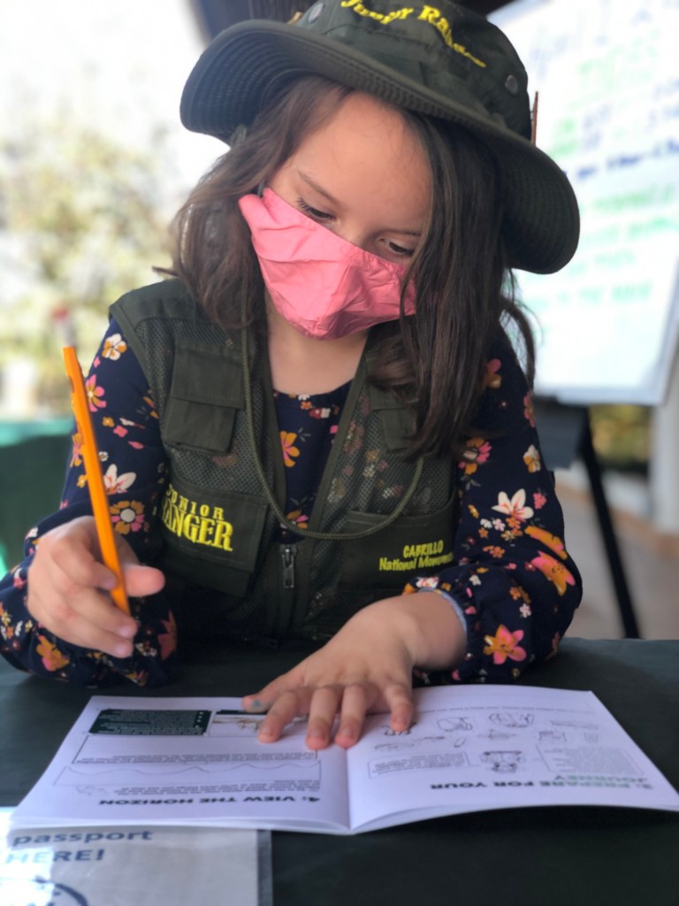A girl with a pink face mask, junior ranger hat and vest, fills out her Junior Ranger booklet