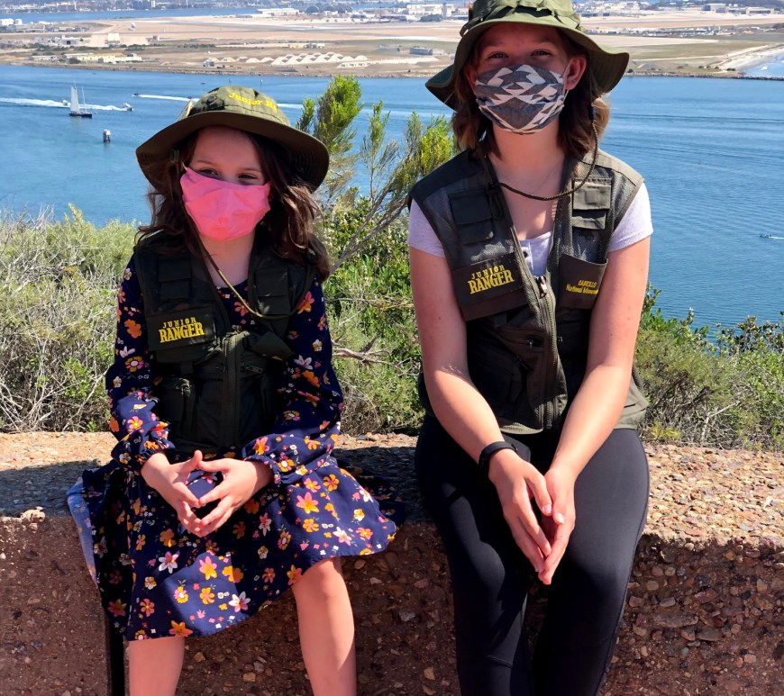 Two girls wear thei Junor Ranger hats and vests, sit on a stone wall with the blue water of San Diego Bay behind them.