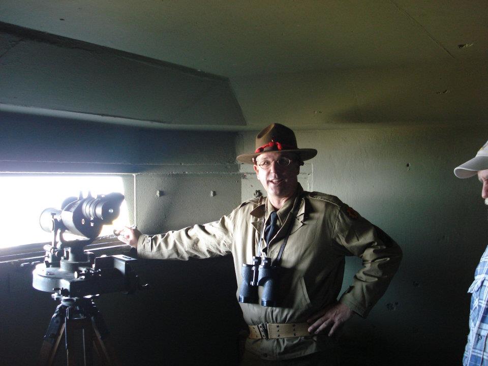 A man in World War II uniform stands next to telescope.