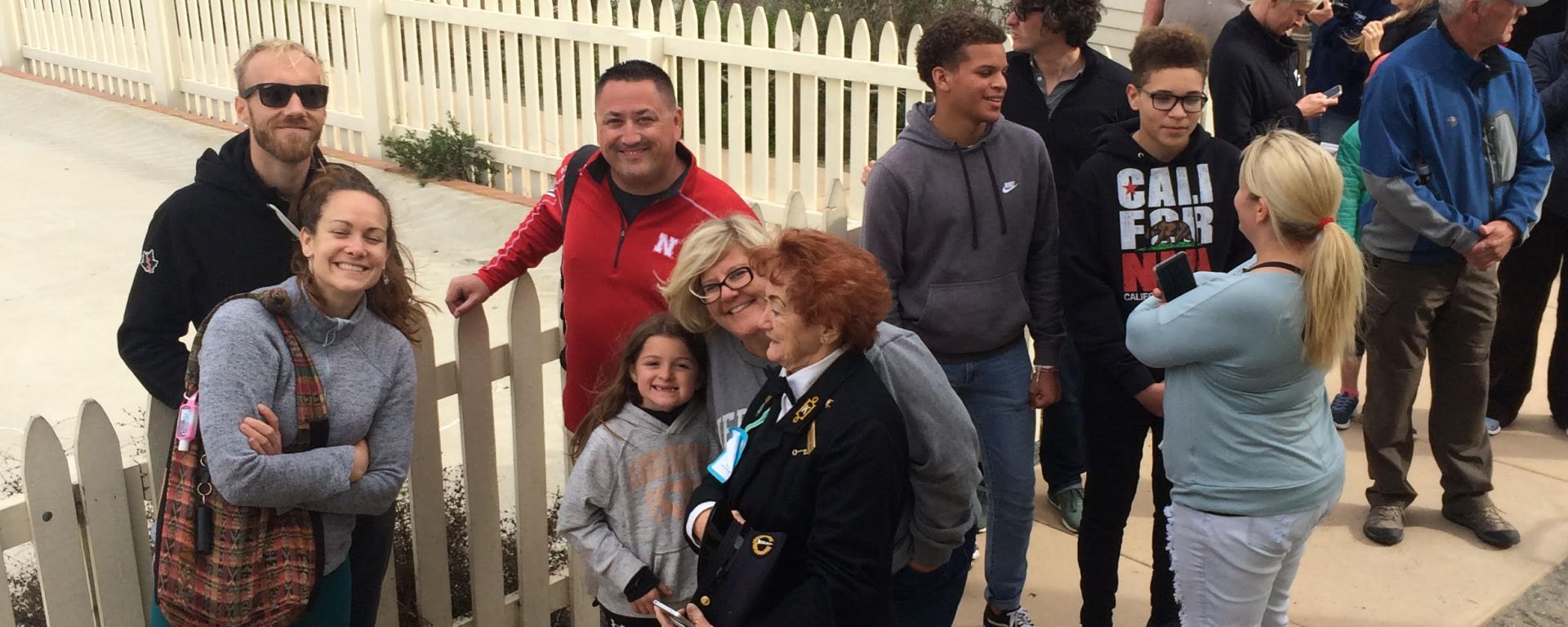 People stand in line along a concrete pathway. A white building and the ocean are in the background.