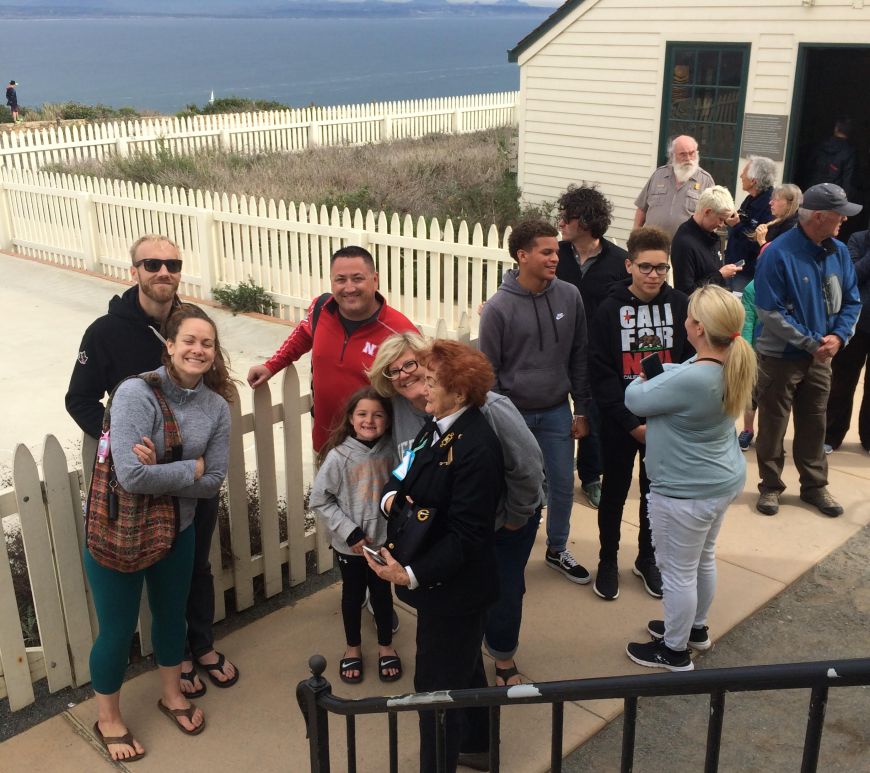 People stand in line along a concrete pathway. A white building and the ocean are in the background.