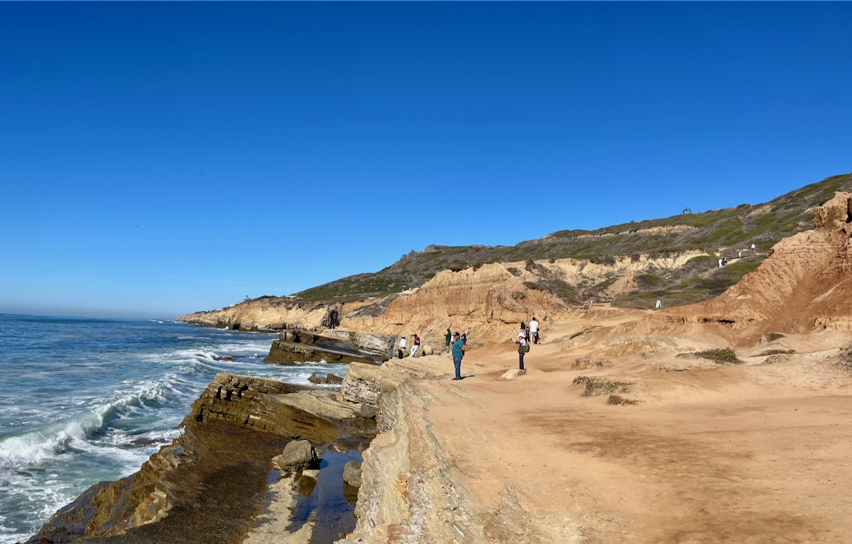 People standing along a tan rocky shoreline.