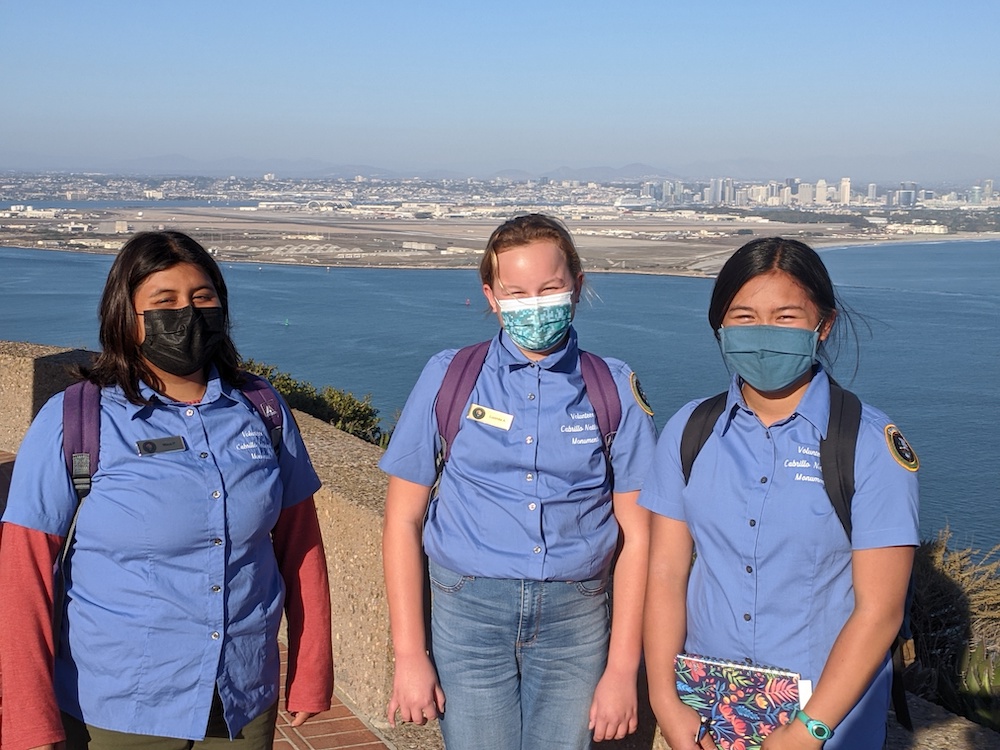Three girls in blue shirts, face masks and backpacks stand with city skyline in background