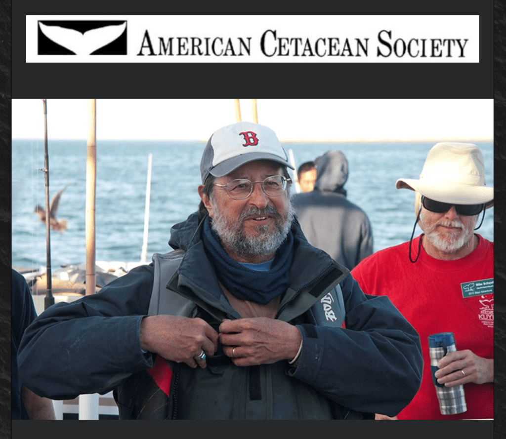 Man with glasses and baseball hat standing with ocean in background