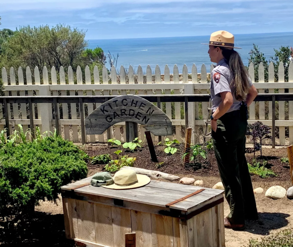 A woman wearing a ranger hat stands in a vegetable garden. The ocean is in the distance.