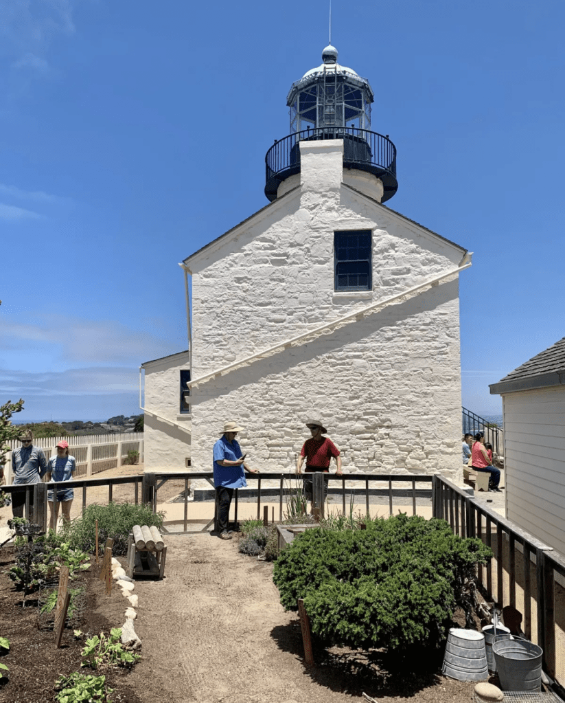 A garden surrounded by a wooden picket fence. A white lighthouse is in the background.
