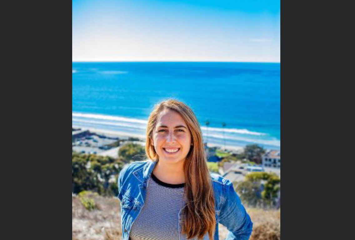 A woman with long hair wears a blue jacket and white pants. The ocean is in the background.