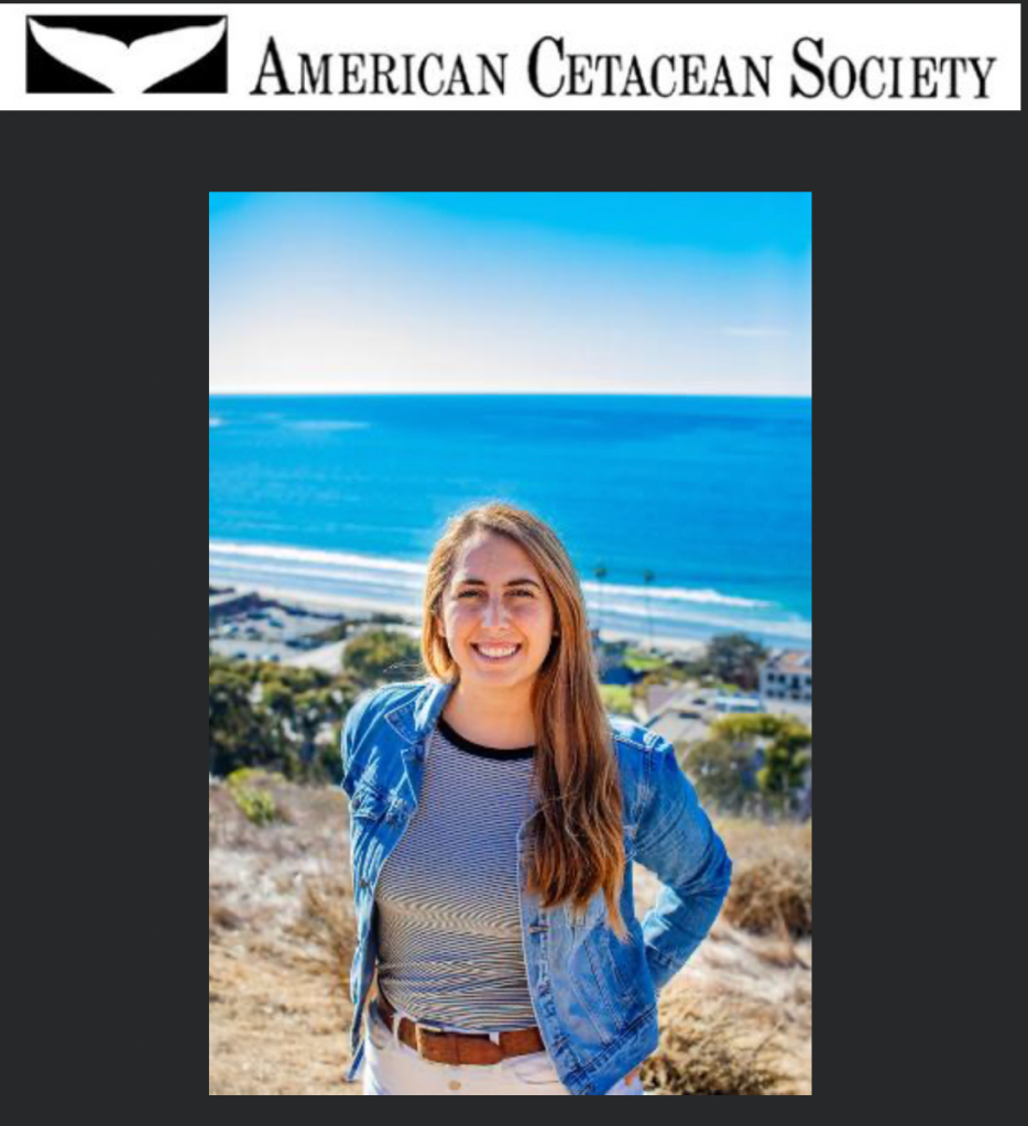 A woman with long hair wears a blue jacket and white pants. The ocean is in the background.