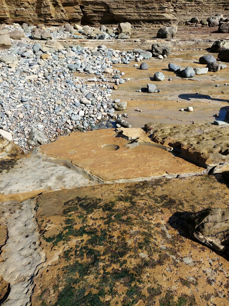 A pile of rocks near a flat slab of rock. A circular hole the size of a basketball is in the middle of the flat rock.