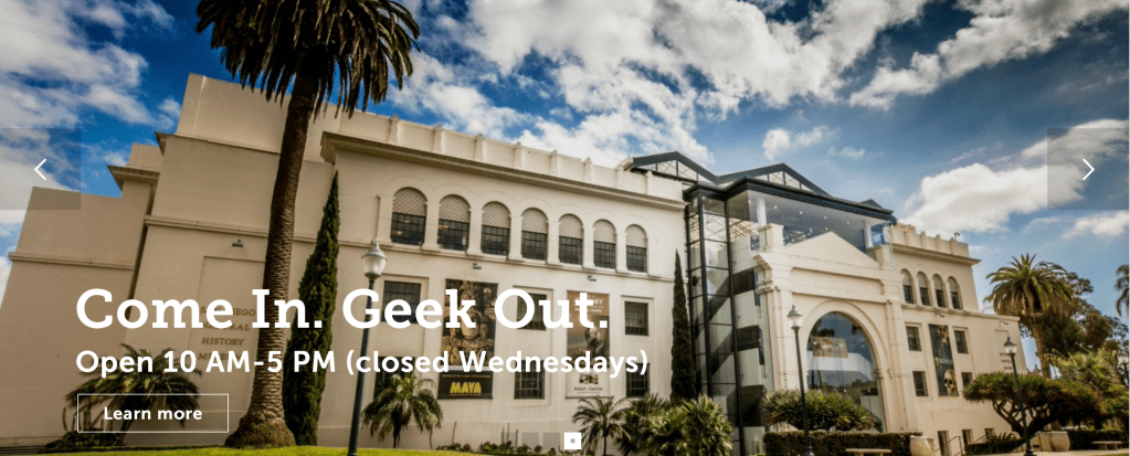 A white multi story building with white puffy clouds among a blue sky. Text reads Come In. Geek Out.