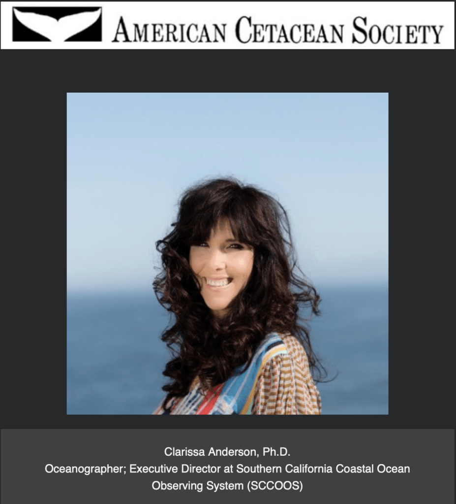 A woman smiles to the camera. The ocean is in the background.