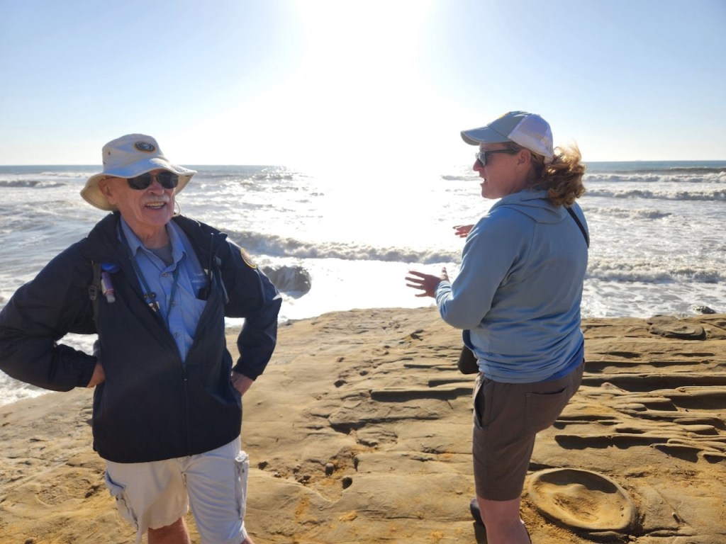 A man and a woman are talking along a rocky beach. The ocean is in the background.