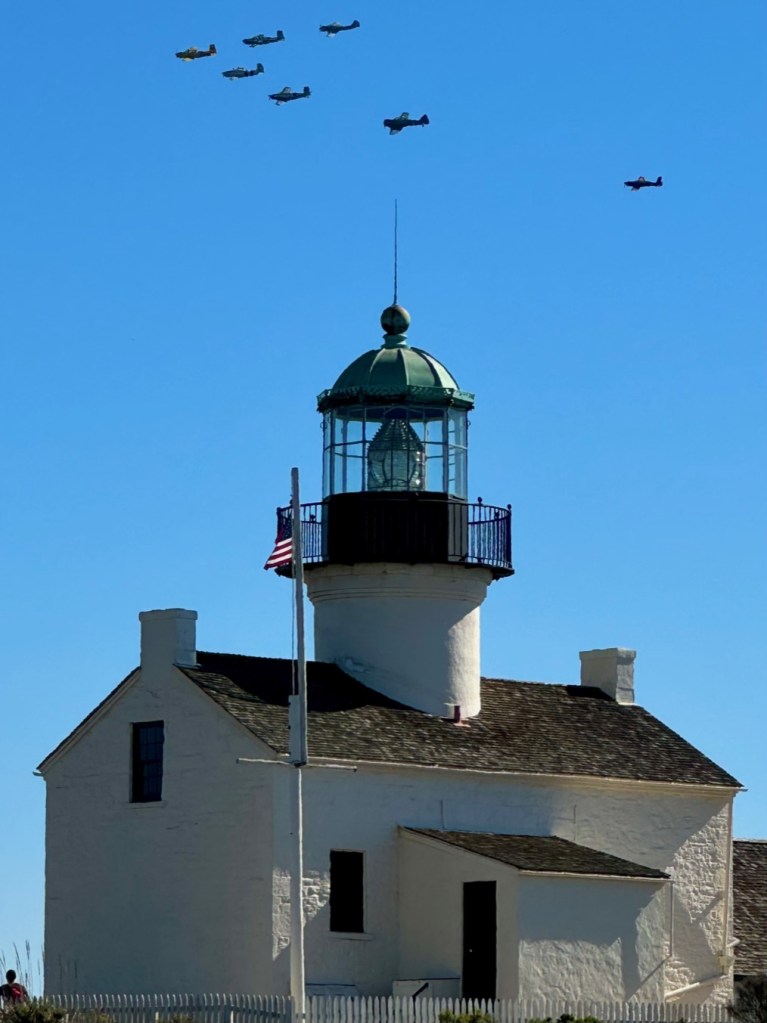 Vintage airplanes fly over a lighthouse with a blue sky in the background.