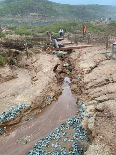 The picture shows a washed out hiking trail. The trail has reddish-brown soil and a trickle of water running through it. Piles of blue-gray stones are scattered at the bottom of the gully, possibly placed there to prevent further erosion. In the background, there are people walking on an undamaged part of the path, and the landscape is hilly with green vegetation.
