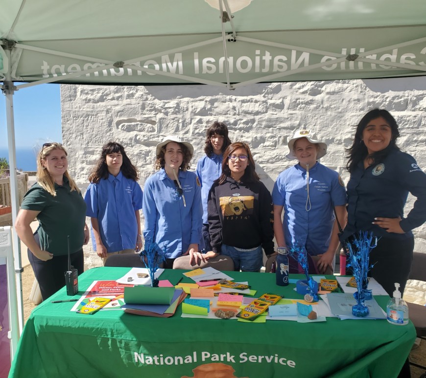 Five youth conservation and environmental stewardship apprentices and two park staff stand behind a table covered in various colored sticky notes and writing utensils, posing for a photo.