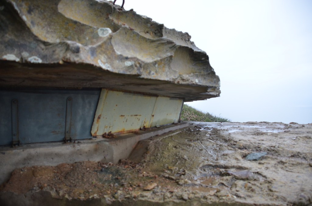 The picture shows a close-up view of a weathered concrete structure with visible signs of erosion and decay. The top part of the structure has a rough, uneven surface with patches of lichen or moss. Below this, there are rusted metal sheets, possibly reinforcements, that are exposed and bending downwards due to deterioration. The ground in front of the structure is muddy with traces of water, indicating recent rain or proximity to a water body.