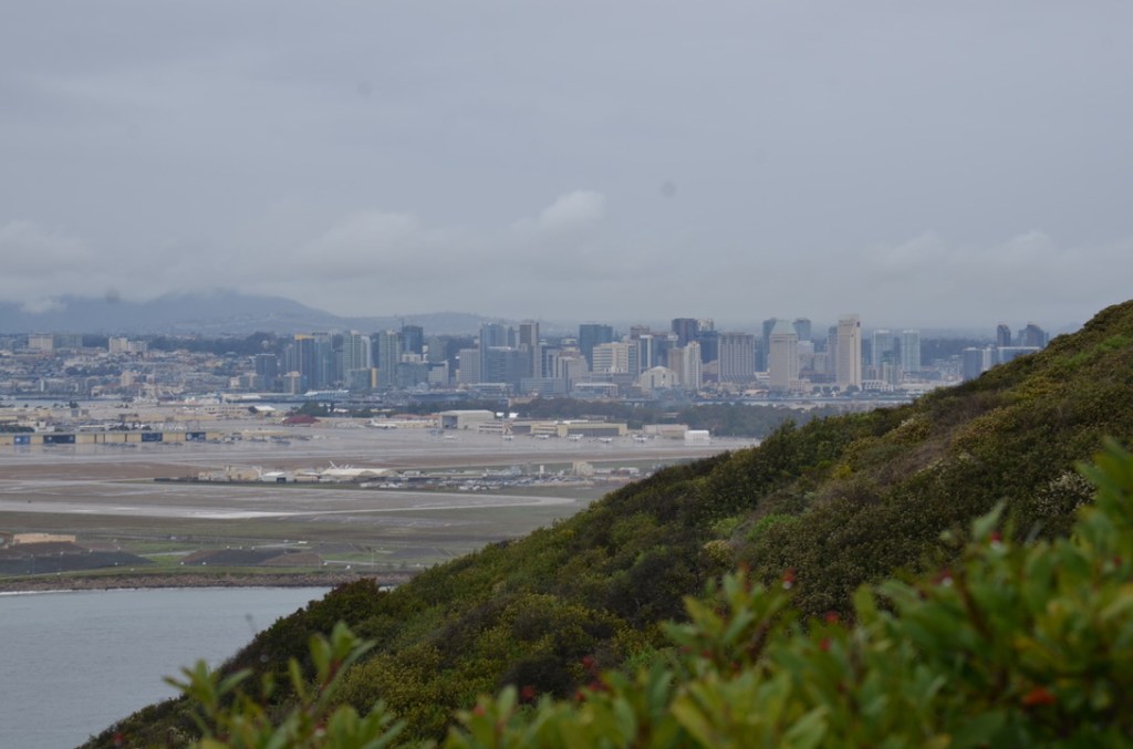 The picture shows a cityscape viewed from a high vantage point. In the foreground, there is a hill covered with green shrubbery. Beyond the hill, an airport with multiple runways and buildings is visible. The middle ground is dominated by the airport's expanse. In the background, a dense collection of high-rise buildings forms the city's downtown area. The sky is overcast with clouds hanging low, partially obscuring the tops of the tallest skyscrapers. The overall color palette is muted with greys, blues, and greens, suggesting a cloudy or overcast day.