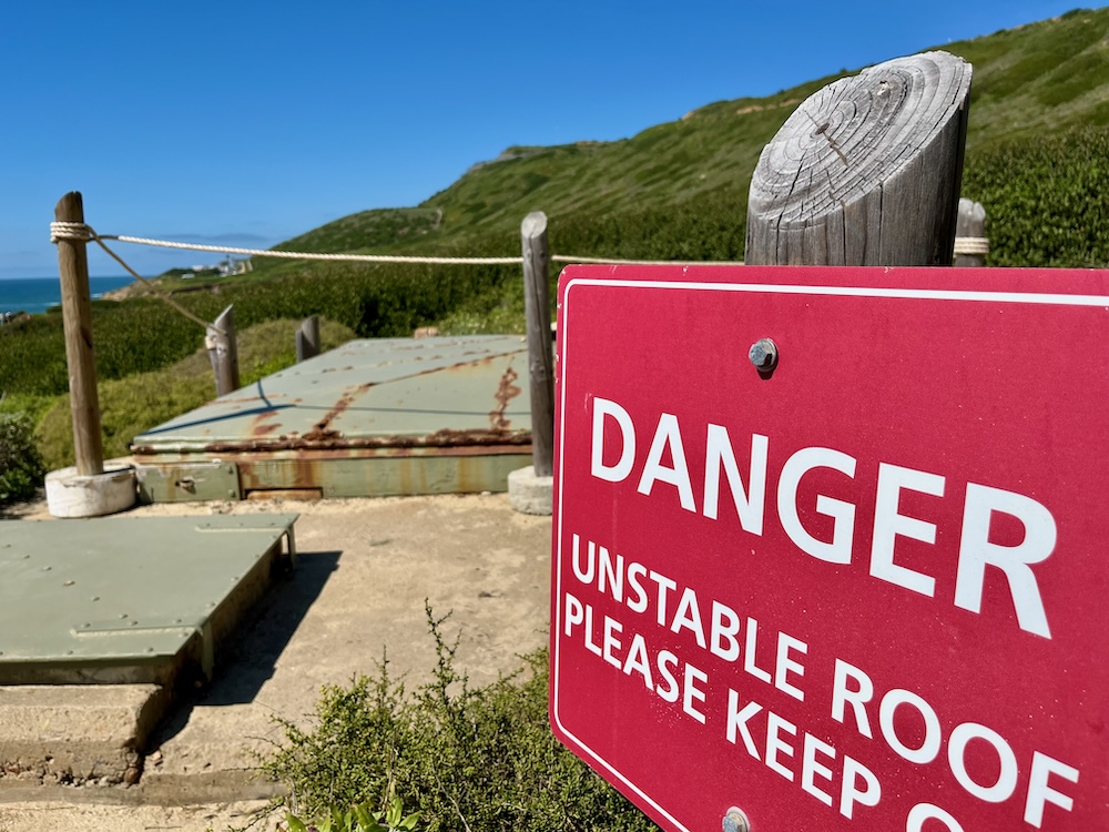 The image shows a bright red warning sign in the foreground that reads "DANGER UNSTABLE ROOF PLEASE KEEP OFF" in bold white letters. Behind the sign, there is a green, rusted metal structure that appears to be the unstable roof mentioned in the sign. The structure is surrounded by a rope barrier attached to wooden posts. In the background, there is a hill covered with greenery and a clear blue sky above.