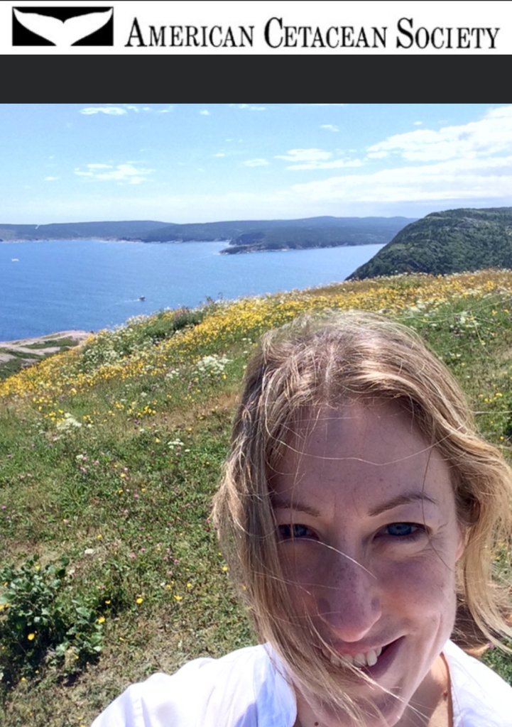 A woman with blond hair on a hillside overlooking a body of water. Yellow flowers are on the hillside.