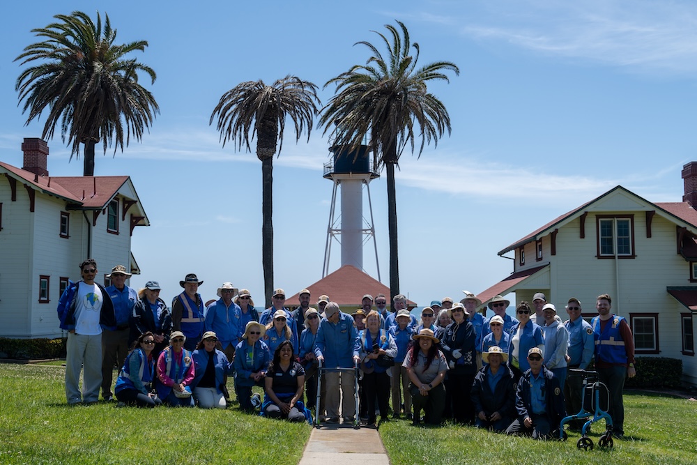 A large group of people in blue shirts stand in front of a lighthouse.