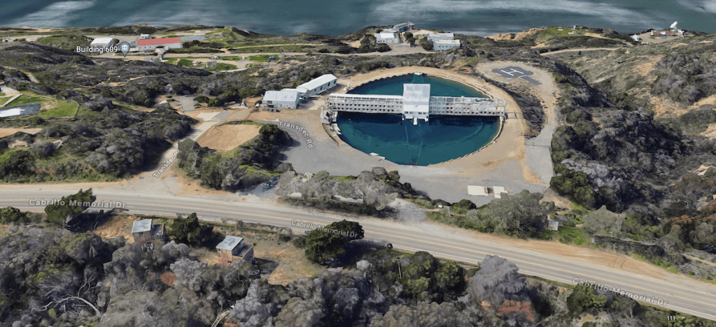 The image shows an aerial view of a coastal area with a large circular structure that appears to be a water tank or reservoir. The structure is surrounded by several buildings and roads. Key features: - The circular water tank is centrally located and has a platform or structure extending over it. - Surrounding the tank are various buildings, including one labeled "Building 609." - A road named "Cabrillo Memorial Dr" runs horizontally across the bottom of the image. - The area is hilly with patches of vegetation and rocky terrain. - In the background, there is a body of water, likely the ocean or a large bay.
