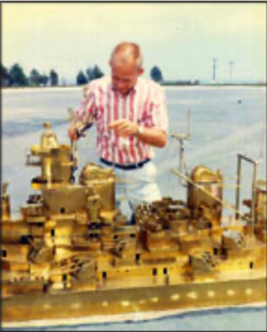 The image shows a man standing outdoors on a paved surface, working on a large, detailed model of a ship. The man is wearing a short-sleeved, red and white striped shirt and light-colored pants. He appears to be adjusting or assembling parts of the ship model, which is predominantly golden in color and has intricate details, including various structures and masts. The background is somewhat blurred, but it shows an open area with some trees and power lines in the distance. The sky is clear, suggesting it is a sunny day.