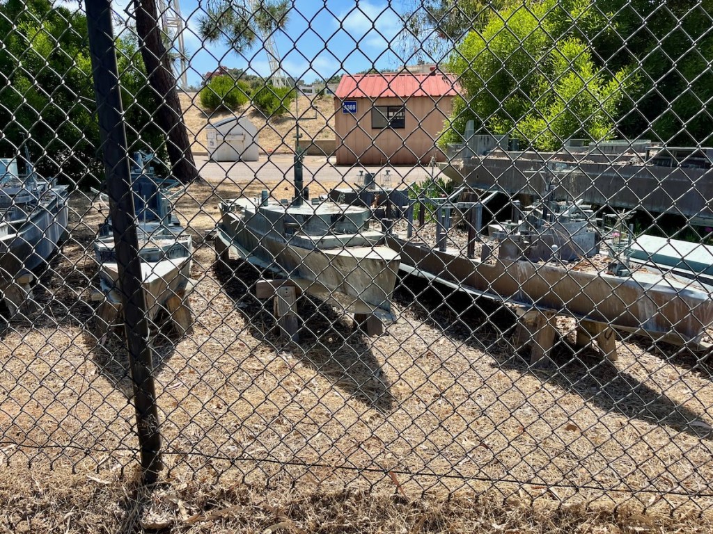 The image shows a fenced area with a chain-link fence in the foreground. Inside the fenced area, there are several metal structures and equipment, possibly related to industrial or utility purposes. The ground is dry and covered with brown grass or dirt. In the background, there is a small building with a red roof and beige walls. The area is surrounded by some greenery, including trees and bushes. The sky is clear and blue, indicating a sunny day.