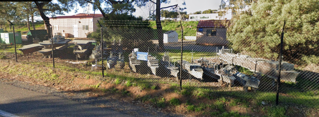 The image shows an outdoor area enclosed by a chain-link fence. Inside the fenced area, there are several large, gray, model ships. The ground inside the fence is a mix of dirt and sparse grass. In the background, there are a few buildings, one of which has a reddish-brown roof and another with a sign that reads "A300." There are also some trees and bushes around the area, providing some greenery. The scene appears to be taken on a sunny day, with shadows indicating the position of the sun.