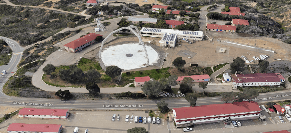 The image shows an aerial view of a complex with several buildings and structures. The most prominent feature is a large circular area in the center, which appears to be a large dish or platform, possibly part of a radar or satellite installation. Surrounding this central structure are multiple buildings with red roofs. The area is bordered by roads, with "Cabrillo Memorial Dr" being the main road visible in the image. The landscape around the complex is hilly and covered with vegetation. There are also parking areas with several vehicles parked. The overall setting appears to be a mix of industrial and natural elements.