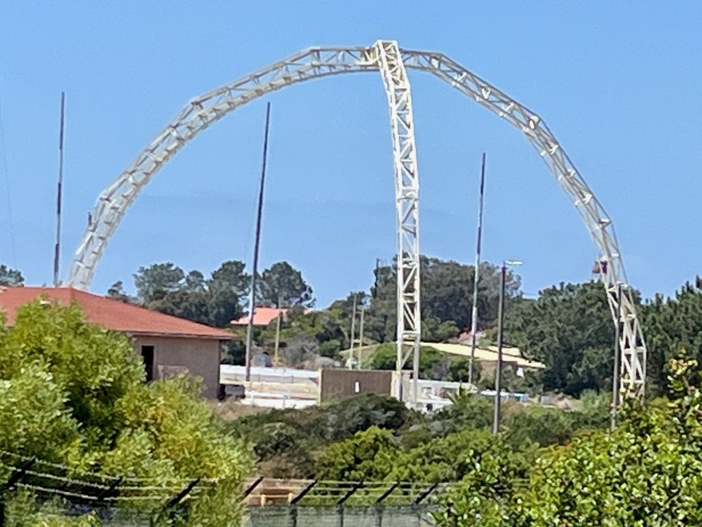 The image shows a large, white, metal arch structure set against a clear blue sky. The arch is tall and has a lattice-like design, with visible cross-bracing. In the foreground, there are green trees and bushes, and a building with a red roof. In the background, there are more trees and some additional buildings. The overall setting appears to be an outdoor area, possibly a park or a facility with open spaces.