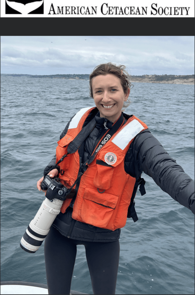 A woman stands on a boat wearing an ornage vest and hold a camera with a long lens.