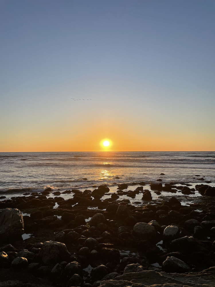 The image shows a serene sunset over the ocean. The sun is low on the horizon, casting a warm golden glow across the sky and reflecting on the water. The sky transitions from a light orange near the horizon to a soft blue higher up. In the foreground, there are dark, rocky formations along the shoreline, with the waves gently lapping against them. A small flock of birds is visible flying in the distance, silhouetted against the sky. The overall scene is peaceful and picturesque.
