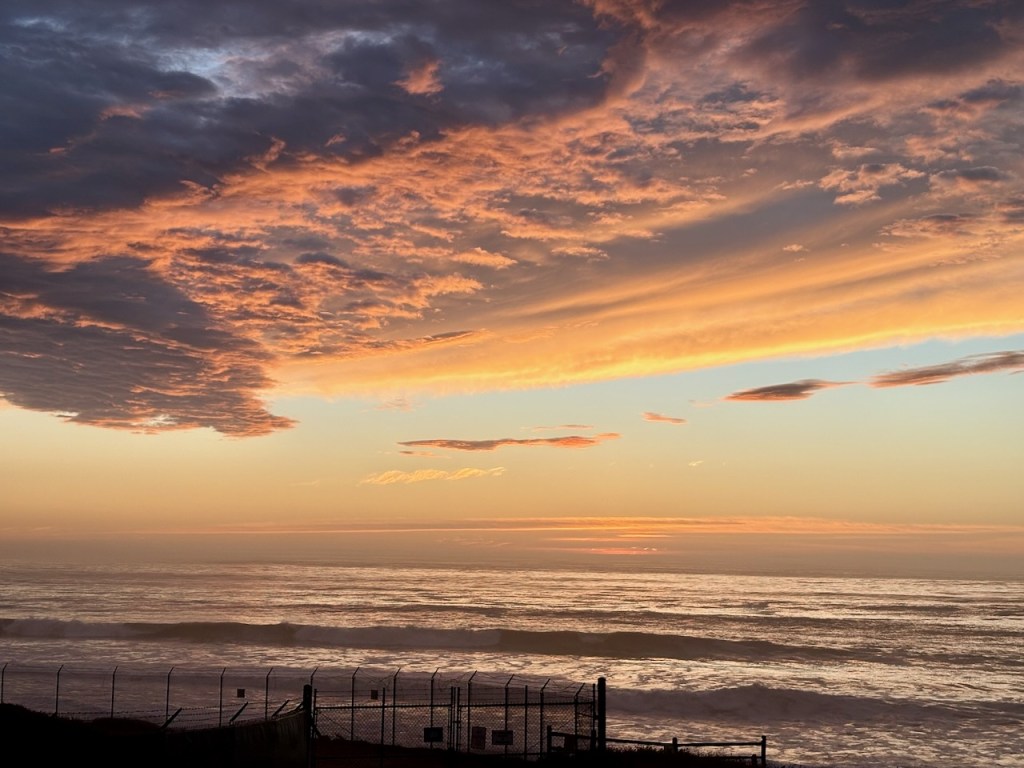 The image shows a beautiful sunset over the ocean. The sky is filled with dramatic clouds that are illuminated in shades of orange, pink, and purple by the setting sun. The ocean below reflects the colors of the sky, creating a serene and picturesque scene. In the foreground, there is a silhouette of a fence with some posts and wires, adding a contrasting element to the natural beauty of the sunset and ocean.