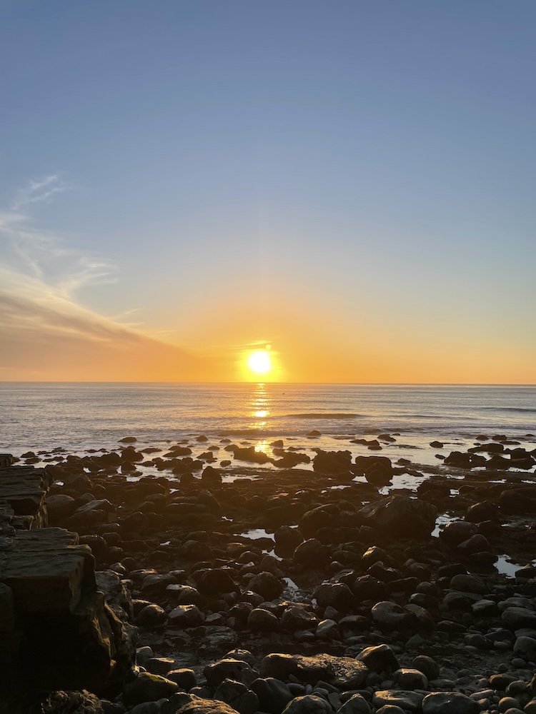 The image shows a serene sunset over the ocean. The sun is low on the horizon, casting a warm golden glow across the sky and reflecting on the water. The sky transitions from a soft orange near the horizon to a clear blue as it extends upward. In the foreground, there is a rocky shoreline with dark, rounded rocks scattered across the scene, partially submerged in shallow water. The overall atmosphere is calm and peaceful.