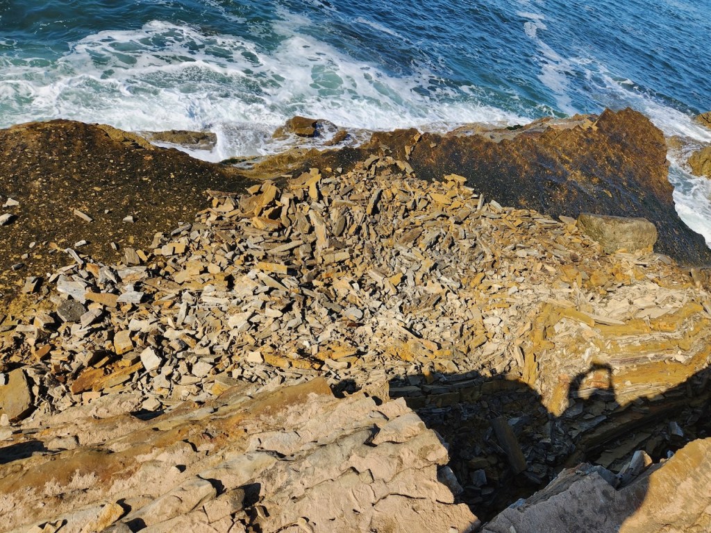 The image shows a rocky coastline with the ocean in the background. The waves are crashing against the rocks, creating white foam. The foreground is filled with various sizes of jagged, layered rocks and stones, mostly in shades of brown and beige. There is a shadow of a person taking the photo, visible on the rocks, with their arms raised, possibly holding a camera or phone. The ocean water is a deep blue, transitioning to lighter shades near the shore where the waves are breaking.