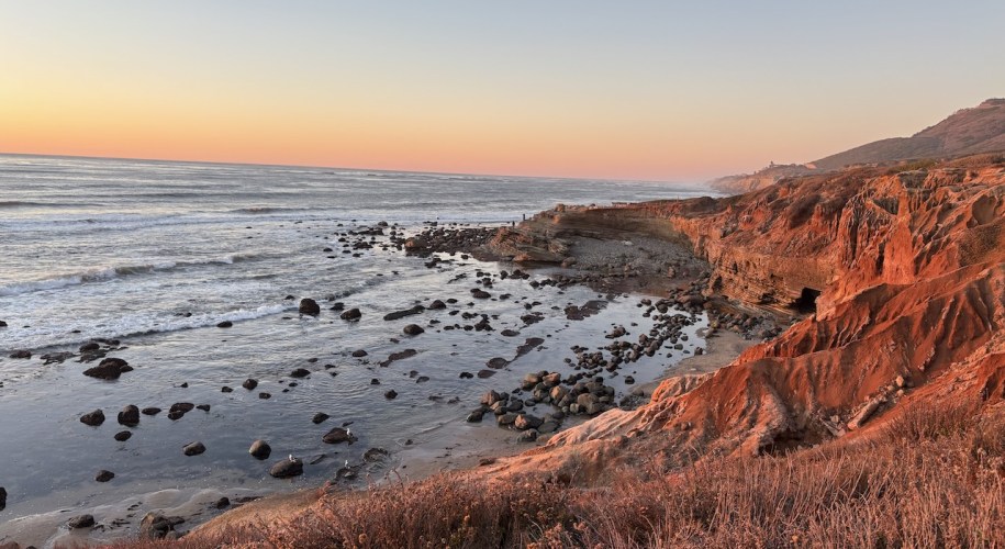 The image shows a scenic coastal landscape during sunset. The foreground features rugged, reddish-brown cliffs with some vegetation. Below the cliffs, there is a rocky shoreline with scattered boulders and small pools of water. The ocean stretches out to the horizon, with gentle waves rolling in. The sky is a gradient of colors, transitioning from a soft orange near the horizon to a pale blue higher up, indicating the sun is setting. The overall atmosphere is calm and serene, with warm tones from the setting sun illuminating the scene.