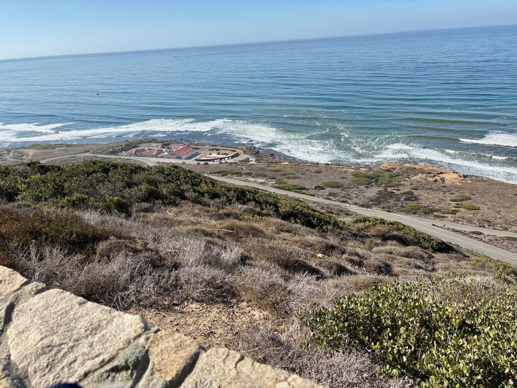 The image shows a scenic view of a coastline. In the foreground, there is a stone wall and some dry, bushy vegetation on a slope. Beyond the slope, there is a road running parallel to the coastline. Further in the distance, there are some buildings with red roofs near the shore. The ocean is visible with gentle waves rolling towards the shore, and the sky above is clear and blue. The overall scene is bright and sunny, suggesting a pleasant day.