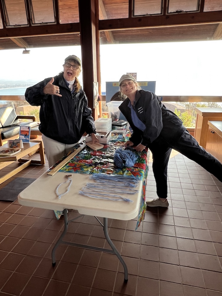 The image shows two women standing indoors next to a table covered with various items. Both women are wearing dark jackets and caps. The woman on the left is making a playful gesture with her hand, resembling a "hang loose" sign, and has a cheerful expression. The woman on the right is leaning slightly forward with one leg extended behind her, also smiling. The table in front of them is covered with a colorful cloth and has several items on it, including a ball of blue yarn, strands of yarn laid out, and some papers or brochures. The setting appears to be a room with large windows, allowing a view of the outdoors, possibly overlooking a body of water or landscape. The floor is tiled, and there are wooden elements in the room's structure.