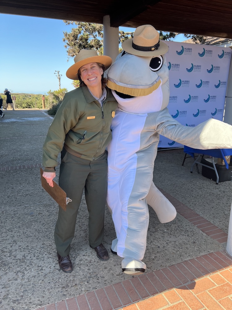 The image shows a person dressed in a park ranger uniform standing next to someone in a dolphin costume. The park ranger is wearing a wide-brimmed hat, a green jacket, and dark pants, and is holding a clipboard. The dolphin costume is light gray and white, with large cartoonish eyes and a big smile. The person in the costume is also wearing a straw hat. They are standing under a covered area with a backdrop that has the "Hubbs SeaWorld Research Institute" logo repeatedly printed on it. The setting appears to be outdoors, with trees and a view of the ocean in the background. Both individuals are smiling and posing for the photo.