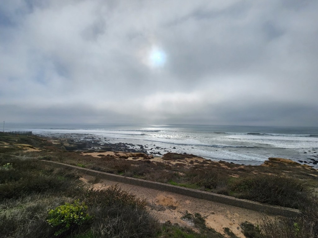 A cloudy sky over a rocky coastline with waves gently rolling onto the shore. The sun is partially visible through the clouds, creating a bright spot in the sky. In the foreground, there is a path running parallel to the coastline, surrounded by low vegetation and shrubs. The overall atmosphere is calm and serene.