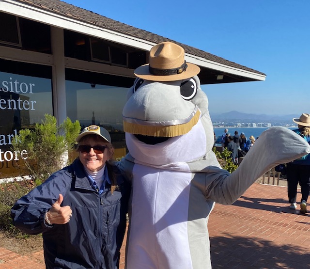 A person wearing a navy blue jacket and a cap is standing next to a person in a large dolphin costume. The dolphin costume is gray and white, with a big smile and large eyes, and is wearing a ranger-style hat. The person in the jacket is giving a thumbs-up gesture. They are outside, in front of a building with large windows. The windows have text on them, partially visible, reading "Visitor Center" and "Park Store." In the background, there is a view of the ocean and distant mountains, with a clear blue sky. Other people are visible in the distance, some also wearing ranger-style hats.