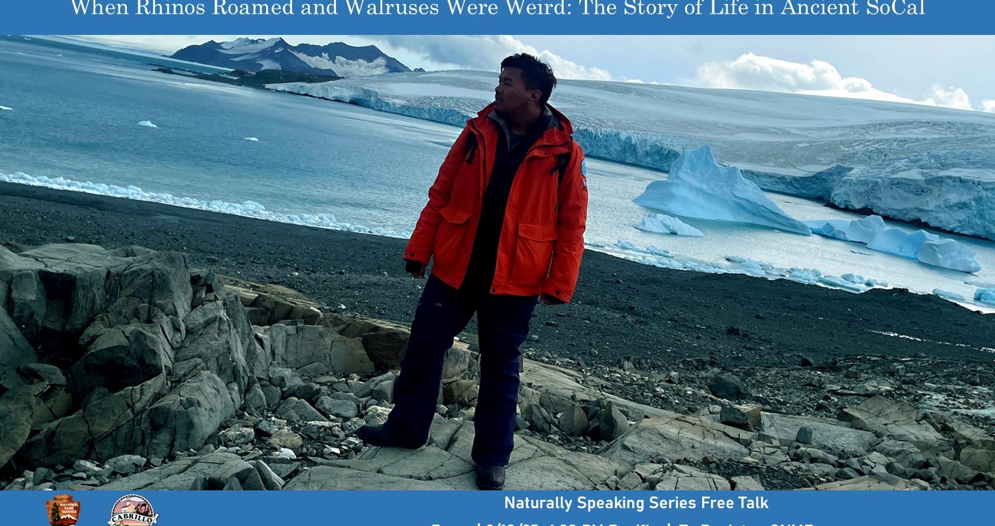 A man in an orange snow jacket and blue snow pants stands on a tumble of gray rocks and looks into the distance with snowy and icy scenery behind him. Text banners above and below the image advertise the event.