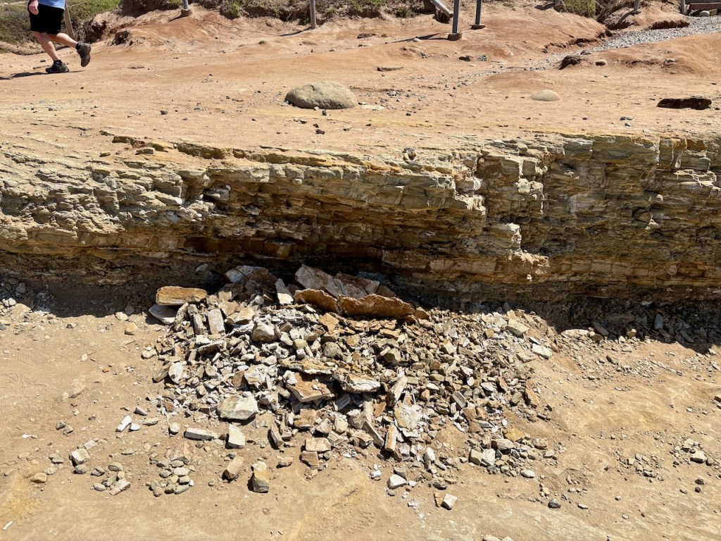 A rocky landscape with a prominent horizontal rock formation. The rock formation appears layered and weathered, with a pile of broken rocks and debris at its base. The ground is sandy and uneven, with scattered small stones. In the background, a person wearing shorts and hiking boots is walking along a path. The area has a dry, arid appearance with sparse vegetation.