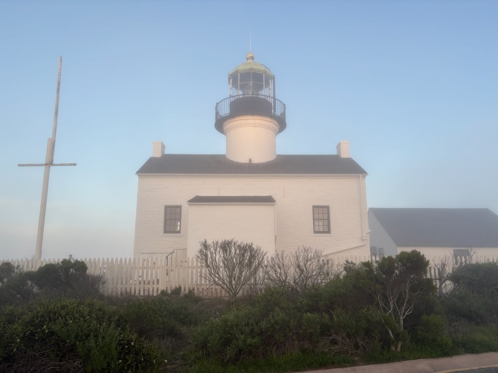 A white lighthouse with a black railing around the top sits under a clear blue sky. The building has a simple, rectangular shape with a pitched roof and two small chimneys on either end. In front of the lighthouse, there is a white picket fence and some low, bushy greenery. To the left of the lighthouse, a tall, white flagpole stands without a flag. The scene has a serene and slightly foggy atmosphere.