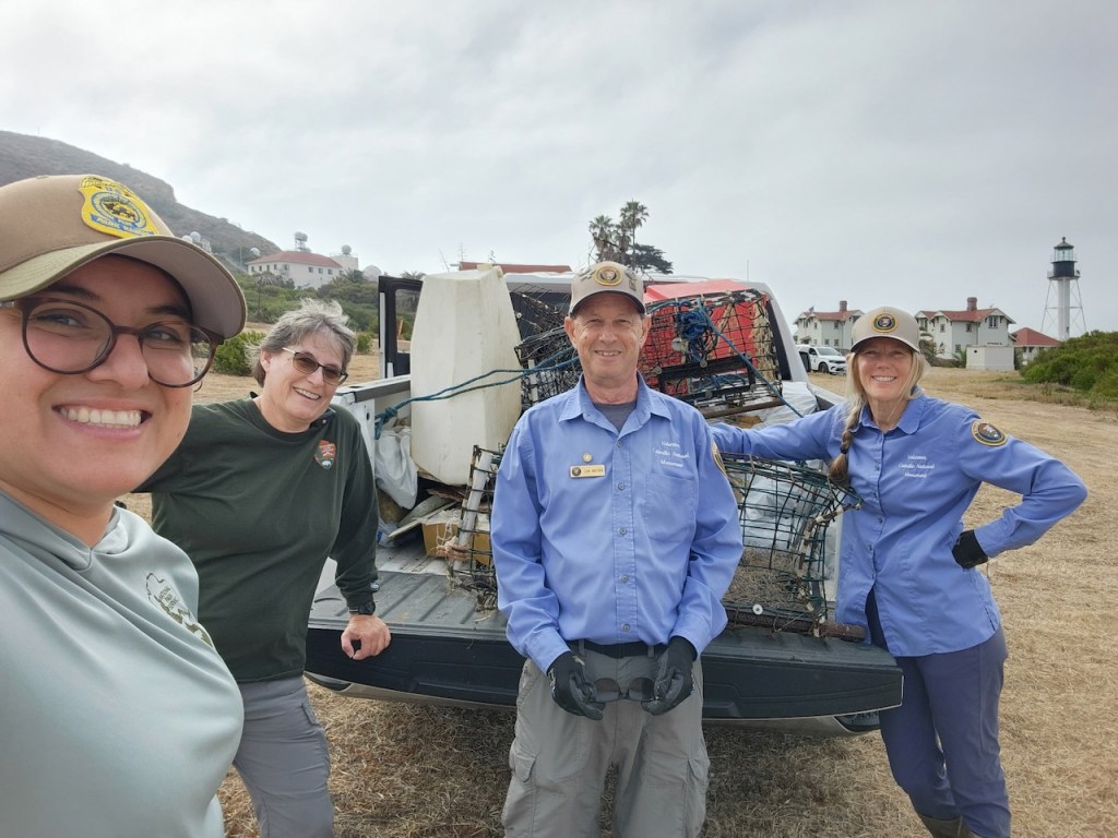 Group of four people standing outdoors in front of a pickup truck loaded with metal cages and equipment. Three women and one man are smiling at the camera. Two women and the man are wearing matching blue shirts and tan hats with a circular patch, while the other woman is in a dark green shirt with a National Park Service badge. The background shows grassy land, a few white buildings with red roofs, and a lighthouse. The sky is overcast, and the group appears to be involved in some outdoor or conservation work.