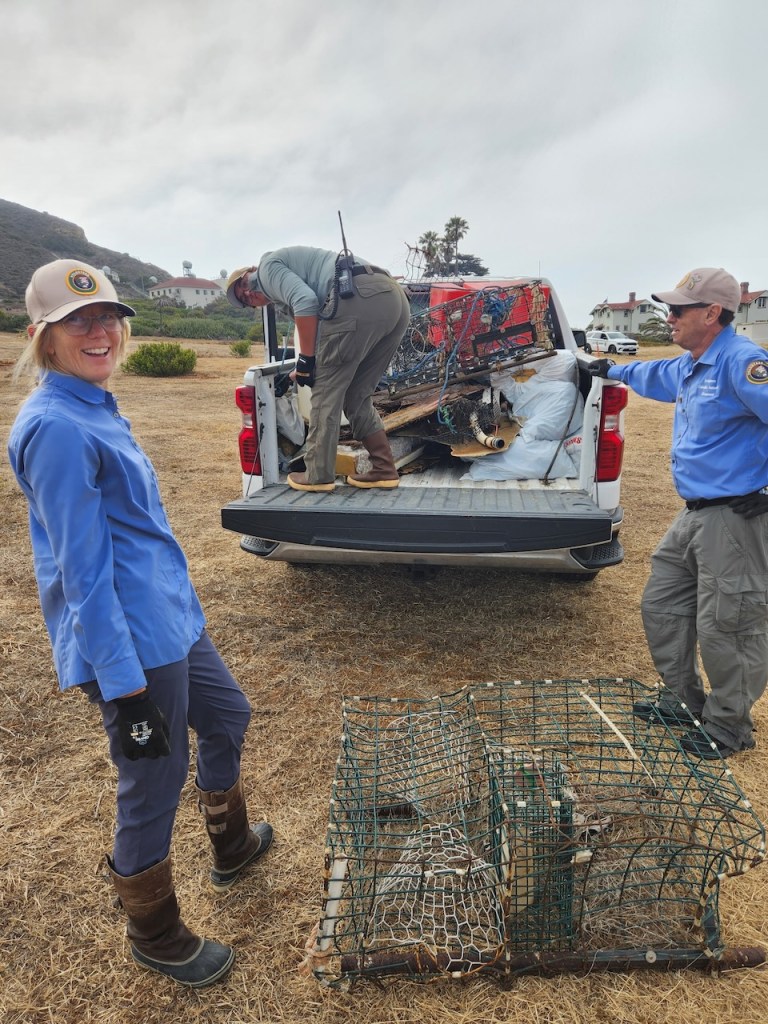 Three people in outdoor uniforms and hats standing near the back of a white pickup truck. The truck bed is filled with collected debris, including metal cages, wood, and bags. One person is standing in the truck bed organizing the items, while the other two stand on the ground, one smiling at the camera and the other holding onto the truck. In front of them on the dry grass are two large, rusty wire cages. The setting appears to be a rural or coastal area with some buildings and palm trees in the background. The sky is cloudy.