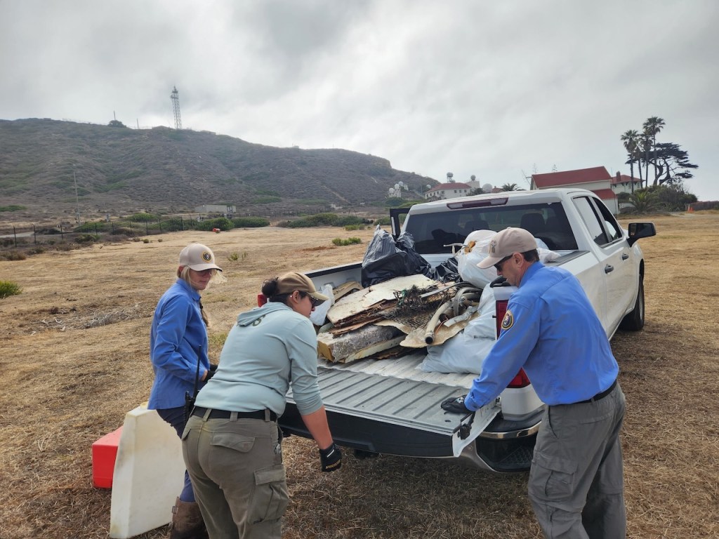 Three people unloading debris and trash from the back of a white pickup truck. The individuals are wearing uniforms with long sleeves, gloves, and caps, suggesting they are part of an organized cleanup effort. The truck bed is filled with various items, including large pieces of debris, black trash bags, and what looks like broken pieces of wood or metal. The scene is set in a dry, grassy field with hills and a lighthouse or tall structure visible in the background. There are a few buildings and palm trees nearby, and the sky is overcast.