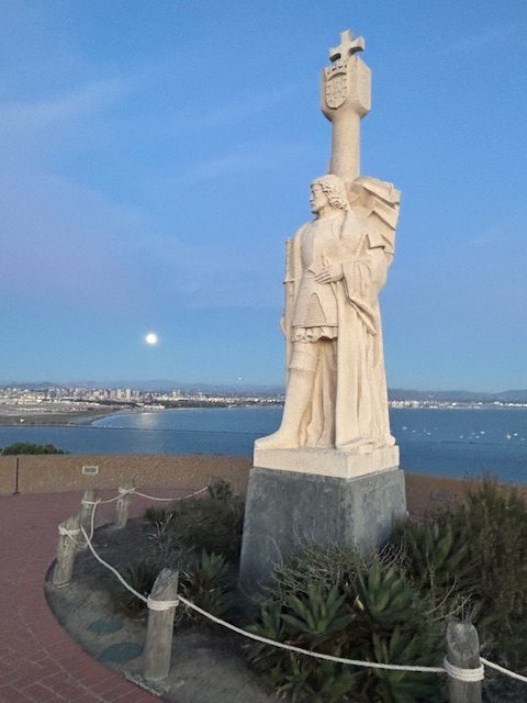 Stone statue of a man in old attire with a cross behind, overlooking water and a city at dusk.