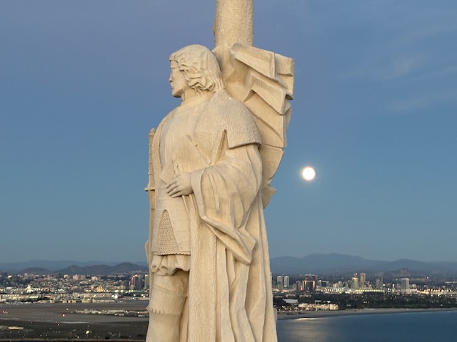 Stone statue in medieval attire overlooking a cityscape at dusk with a full moon behind it.